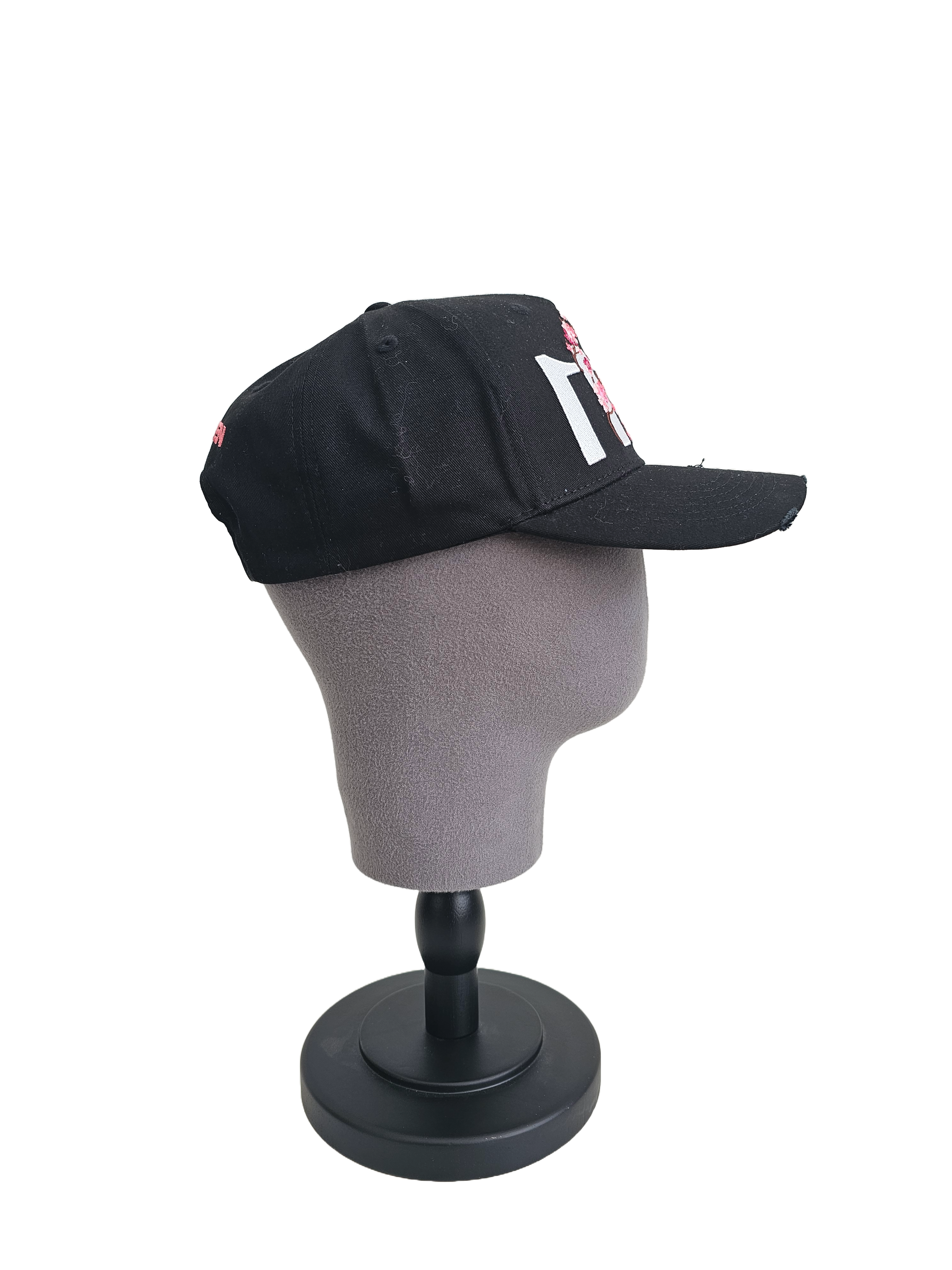 A left sided view of a black snapback baseball cap on a mannequin head with hints of distressed texture and with embroidered pink floral blossom patterns between the 11.11 logo on a white backdrop.