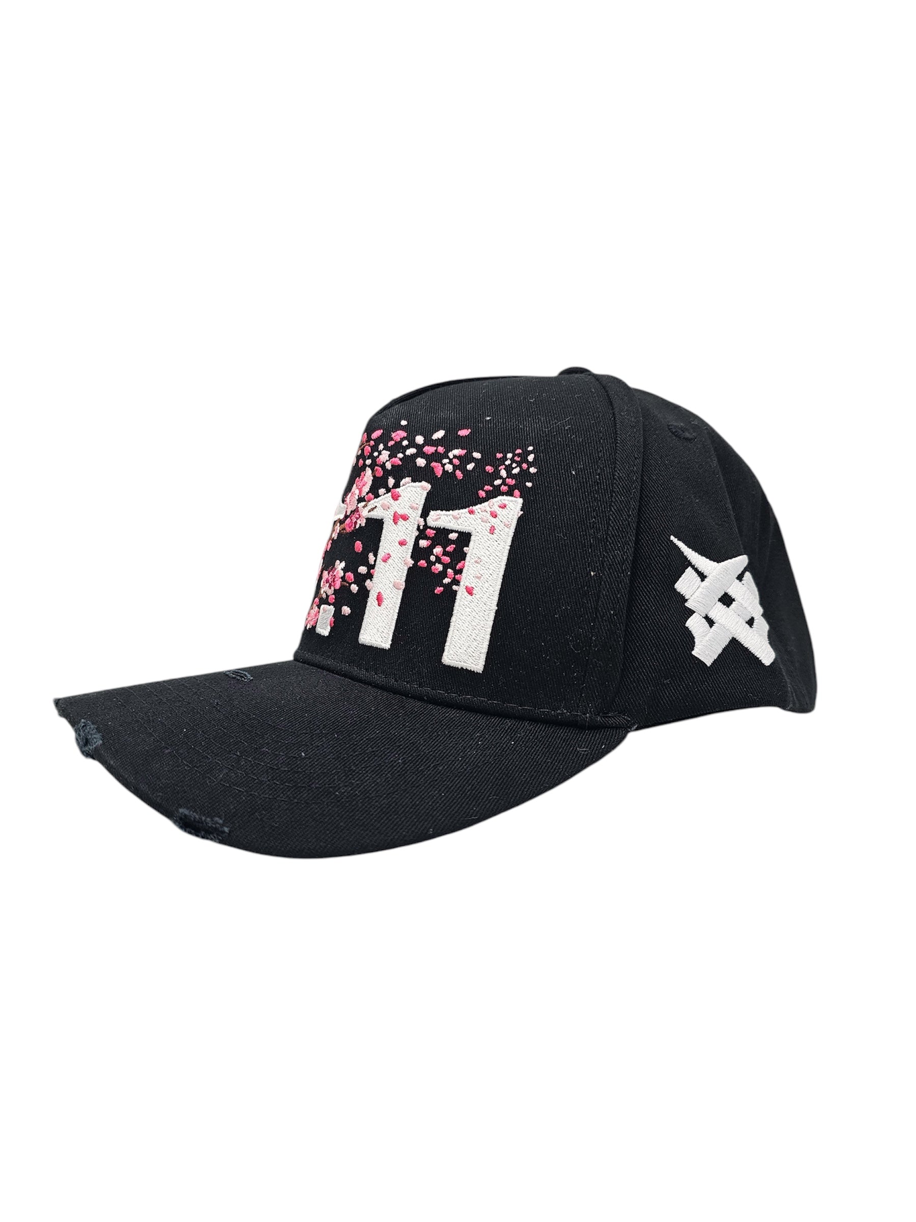 A right angled view of a black snapback baseball cap with hints of distressed texture and with embroidered pink floral blossom patterns between the 11.11 logo on a white backdrop. The cap also reveals a white embroidered 11.11 symbol logo on the right side of the cap.